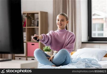 television, mass media and people concept - happy smiling girl with remote control watching tv at home. happy smiling girl watching tv at home