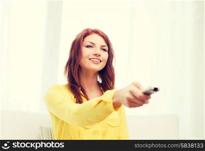 television, relax, home and happiness concept - smiling teenage girl sitting on couch with tv remote control at home