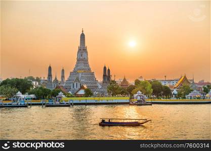Temple of Dawn or Wat Arun and boats on Chao Phraya River at sunset with shining sun. Bangkok, Thailand