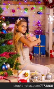 Ten-year-old girl sits at the Christmas tree and hugs a gift