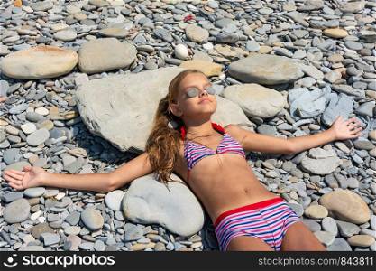 Ten-year-old girl sunbathes on a rocky beach, putting pebbles on her eyes