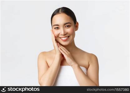 Tender beautiful asian girl in towel standing over white background in bathroom, apply skincare products, cleansing gel for face, touching skin and smiling pleased, white background.. Tender beautiful asian girl in towel standing over white background in bathroom, apply skincare products, cleansing gel for face, touching skin and smiling pleased, white background