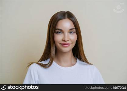 Tender feminine woman looks with admiration and affection, wears white t shirt, has makeup, healthy skin, dark hair, looks at something wonderful, isolated over beige studio wall. Beauty, cosmetology