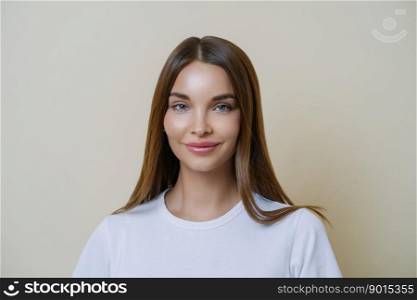 Tender feminine woman looks with admiration and affection, wears white t shirt, has makeup, healthy skin, dark hair, looks at something wonderful, isolated over beige studio wall. Beauty, cosmetology