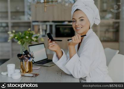 Tender lovely woman with healthy skin holds small mirror and cosmetic brush applies powder on face smiles toothily wears soft white bathrobe and towel poses against home interior. Beauty concept