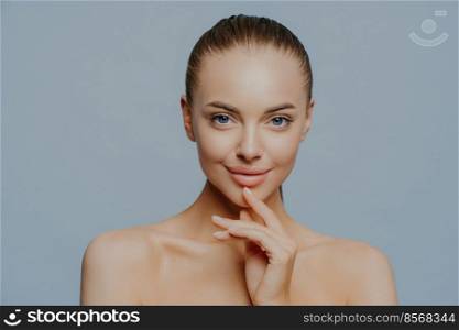 Tender sensual woman with pure healthy skin, has natural magnetic look, feels beautiful and attractive, stands shirtless against blue background, enjoys herself after spa procedures. Beauty care