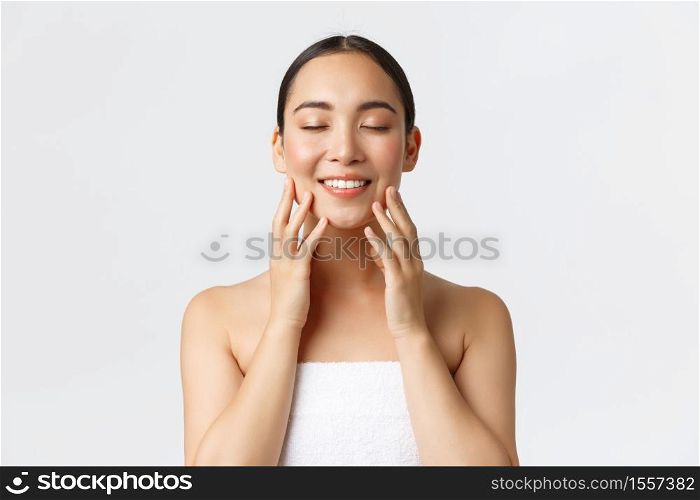 Tender smiling beautiful asian girl attend spa salon, standing in towel, close eyse and touching soft clean skin, satisfied with cream or cleansing gel results, standing white background.. Tender smiling beautiful asian girl attend spa salon, standing in towel, close eyse and touching soft clean skin, satisfied with cream or cleansing gel results, standing white background