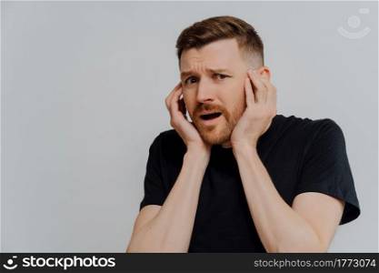 Terrible news. Young shocked man in black shirt holding head in hands and being afraid and scared, guy with horrified facial expression posing against grey background. Fear and fright concept. Shocked scared man looking at camera with frightened face expression
