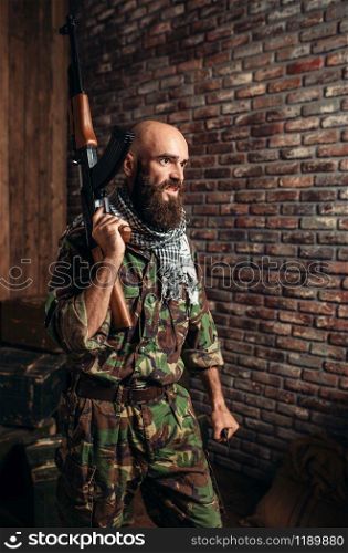 Terrorist in uniform holds kalashnikov rifle in hands, male mojahed with weapon. Terrorism and terror, soldier in khaki camouflage, barrels of fuel or chemicals on background. Terrorist in uniform holds kalashnikov rifle