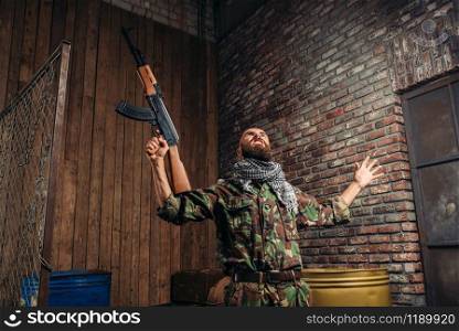 Terrorist in uniform with kalashnikov rifle raised his hands up, male mujahedin with weapon. Terrorism and terror, soldier in khaki camouflage, barrels of fuel or chemicals on background. Terrorist with kalashnikov rifle raised hands up