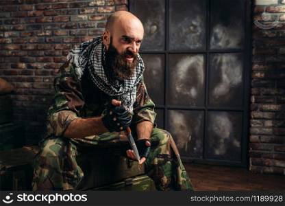 Terrorist in uniform with knife in hands sitting on boxes of ammunition. Terrorism and terror, soldier in camouflage in weapon arsenal, barrels of fuel or chemicals on background. Terrorist with knife sits on boxes of ammunition