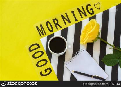 Text Good morning, Cup of coffee, yellow rose and empty notebook for text on stylish black and white napkin on yellow background. Minimal style workplace. Concept good morning. Flat lay Top view Copy space.. Text Good morning, coffee, yellow rose, notebook for text on stylish black and white napkin on yellow background. Minimal style workplace. Concept good morning