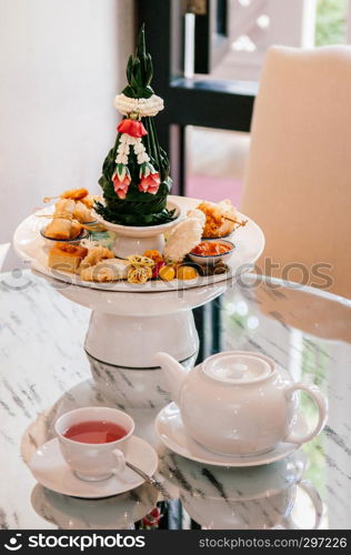 Thai assorted traditional dessert snack, curry puff, rice cracker, custard, spring rolls etc. on white ceramic pedestal tray with tea set. close up shot