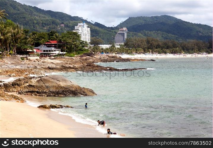 Thai children are swimming in the wild beach. In the background, Patong beach and modern hotels. Editorial only.