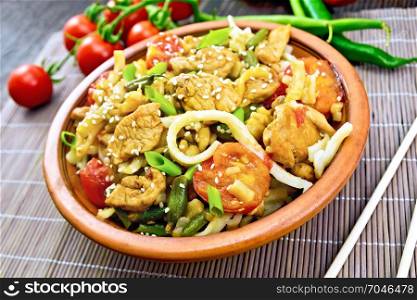 Thai noodles wok with chicken meat, tomatoes, soy sauce and green beans sprinkled with sesame seeds in a clay dish on a bamboo napkin on a wooden board background
