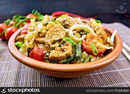 Thai noodles wok with chicken meat, tomatoes, soy sauce and green beans sprinkled with sesame seeds in a clay dish on a bamboo napkin against a dark wooden board