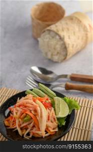 Thai papaya salad in a black plate with Sticky rice, Spoon, and fork.