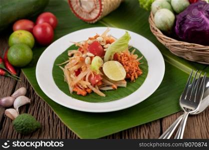Thai papaya salad in a white plate on banana leaves with lime, tomatoes, eggplant, chili, garlic, peppers, salad, and peanuts. 