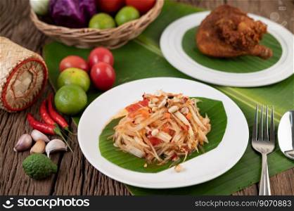 Thai papaya salad in a white plate on banana leaves with lime, tomatoes, eggplant, chili, garlic, peppers, salad, and peanuts. 