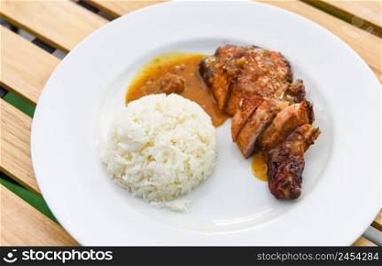Thai rice food, cooked white rice and grilled chicken with sauce on white plate and wooden table background, Spicy bbq chicken legs grilled