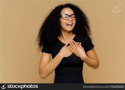 Thankful positive woman smiles happily, makes gratitude gesture, keeps hands on chest, smiles broadly, wears casual t shirt, optical glasses, closes eyes from pleasure, isolated on beige background
