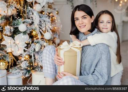 Thankful small female child embraces her mother who gave present, spend wonderful unforgettable time together, celebrate Christmas. Brunette woman and daughter look for present gifts under fir tree