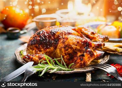 Thanksgiving day dinner table setting with whole roasted turkey or chicken on plate with cutlery , festive lighting and decoration, close up