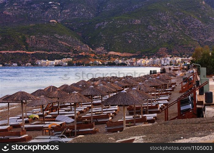Thatched peaks of beach umbrellas with deck chairs on the deserted shore of the sea promenade against the background of evening red twilight light and the rays of the setting sun, mountain ranges in haze and blur with the city of Loutraki in Greece on the horizon.. Thatched peaks of beach umbrellas and wooden deck chairs with mattresses on a deserted promenade among mountains and monasteries in the distance in the rays of the evening setting sun of the city.