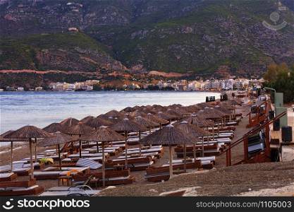 Thatched peaks of beach umbrellas with deck chairs on the deserted shore of the sea promenade against the background of evening red twilight light and the rays of the setting sun, mountain ranges in haze and blur with the city of Loutraki in Greece on the horizon.. Thatched peaks of beach umbrellas and wooden deck chairs with mattresses on a deserted promenade among mountains and monasteries in the distance in the rays of the evening setting sun of the city.