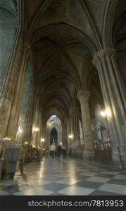 The aisle of the Notre Dame de Paris, with its imposing arches, tiled floor, and many tourists; nicely lit by commemorating candles and the old feel of religion and worship.