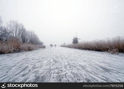 The archetypal Dutch winter on a foggy morning on a frozen canal surrounded by reed, willows, windmills and a handful of ice skaters approaching in the distance