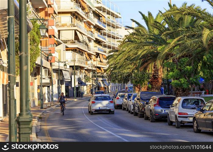 The asphalt road of the street of Loutraki city, which is flooded with sunlight and a teenager rides a bicycle on the road, the sky is reflected in blue light on the roofs of passing cars in the shade of spreading palm trees.. Street landscape of the summer city of Loutraki, Greece, with passing cars and a teenager on a bicycle.