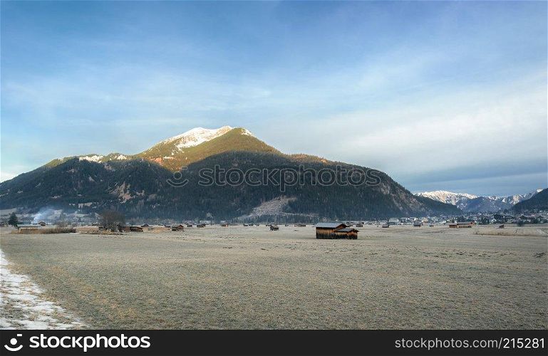 The Austrian Alps mountains, a village, and scattered barns on a meadow with frozen grass, near Ehrwald, Austria, in winter.