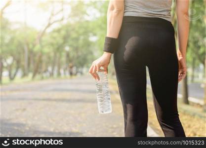 The back of a woman exercising is standing, holding a bottle of water on a tree-lined street with morning sunlight shining, exercise and health concepts.