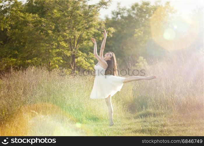 The ballerina doingis arabesk at the meadow In the white dress. At the nature background with the rays of sunset.