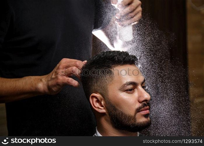 The barber dressed in a black clothes is doing the hair styling to the stylish black-haired man sitting in the armchair in a barbershop