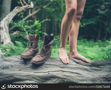 The bare feet of a young woman standing on a fallen tree in the forest with her boots next to her