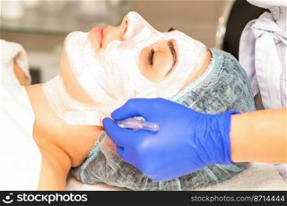 The beautician with brush applies a photochemical and glycolic peeling face mask to the female patient face in the beauty clinic. The beautician with brush applies a photochemical and glycolic peeling face mask to the female patient face in the beauty clinic.