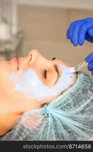 The beautician with brush applies a photochemical and glycolic peeling face mask to the female patient face in the beauty clinic. The beautician with brush applies a photochemical and glycolic peeling face mask to the female patient face in the beauty clinic.