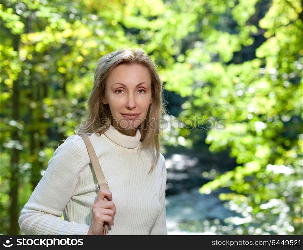 The beautiful woman in a white sweater on the bank of the lake against bright green foliage
