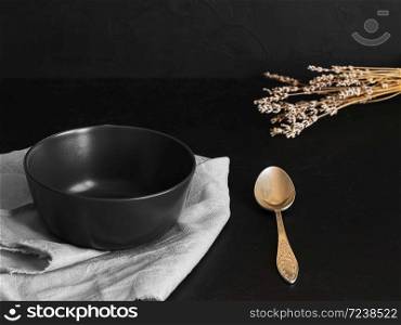 The black bowl is located on a gray napkin. Nearby is a spoon and dry lavender branches. Items are located on a black background. Close-up.