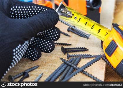 The black screw in hand and is measured by a yellow tape measure with a ruler on a wooden board background. The concept of tools and repair work. Steel screws. Metal screw. The black screw in hand and is measured by a yellow tape measure with a ruler on a wooden board background. The concept of tools and repair work. Steel screws.