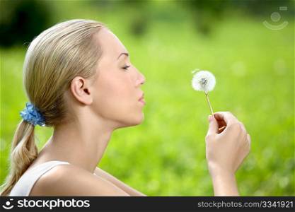 The blonde sits in park on a grass with a dandelion in hands
