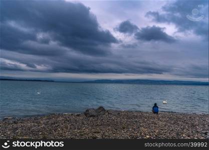The Bodensee lake viewed from Friedrichshafen, Germany, under stormy clouds and a man sitting on its shore, thinking.