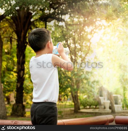 The boy is drinking water from a plastic bottle after exercising While the sun is falling, there is light passing through the trees.