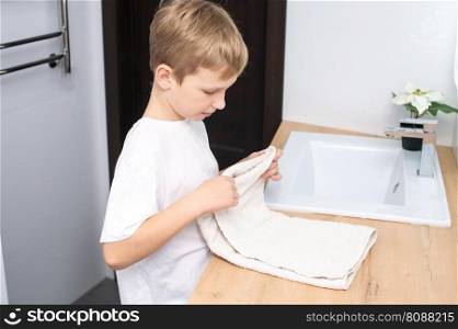The boy is standing in the bathroom near the washbasin and folds a towel. The boy folds a towel and stands in the bathroom near the washbasin
