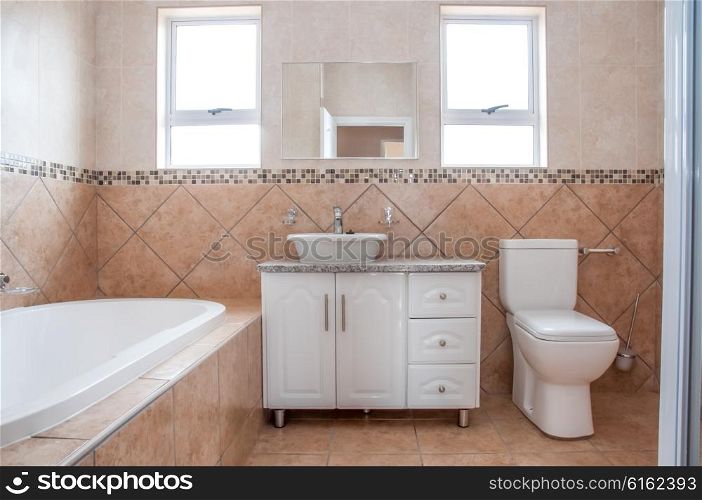 The brand new bathroom of a newly build house, revealing the bath, basin, and the toilet, all in white, in front of brown tiles on the wall.
