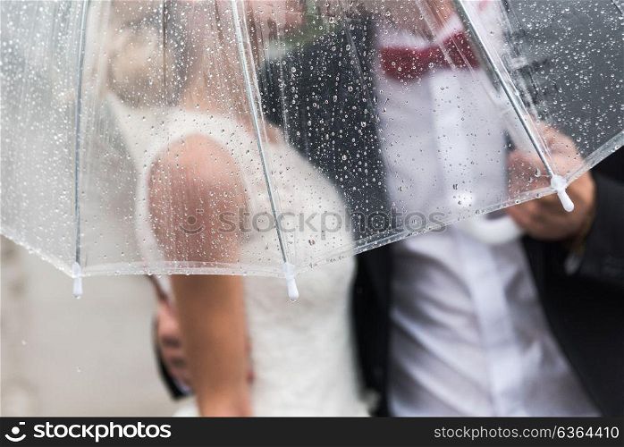 the bride and groom in the rain are covered with a transparent umbrella, rain drops