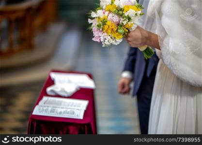 The bride holds a wedding bouquet of beautiful flowers in her hand. The bouquet consists of white, yellow and pink flowers. Marriage registration certificate and grom&rsquo;s hand is In the blur area. Bouquet of flowers in the hand of the bride during the marriage ceremony.