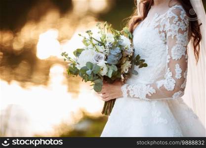 The bride in an elegant wedding dress holds a beautiful bouquet of different flowers and green leaves. Wedding theme.. The bride in an elegant wedding dress holds a beautiful bouquet of different flowers and green leaves. Wedding theme